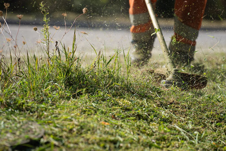A gardener wearing orange trousers and dark boots is using a garden rake to clear overgrown grass and weeds on a lawn. The rake's metal tines are stirring the soil and debris, while grass clippings an