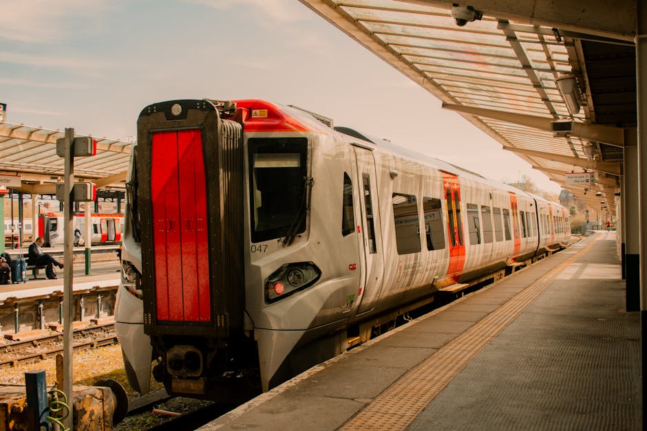 A modern train with a sleek silver exterior and orange accents is parked at a railway station platform under a covered shelter. The train's front features a large, vertically oriented red panel, while